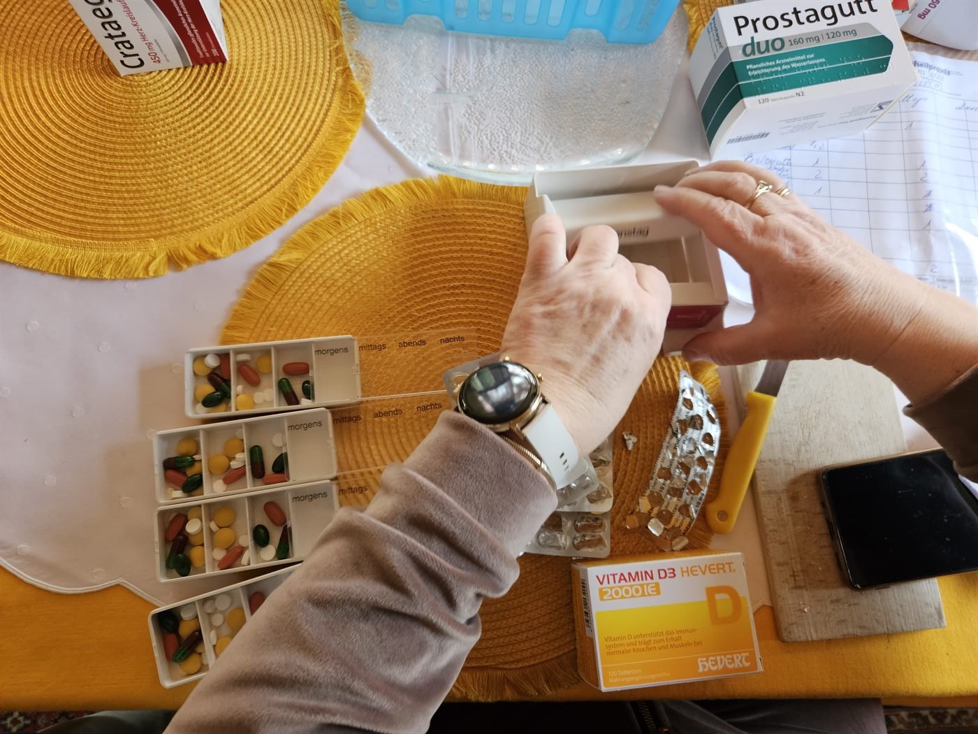 Pflegende Angehörige stellen die Medikamente für einen Wochendispenser mit den Fächern morgens, mittags, abends und nachts – daneben Blister, Arzneimittelpackungen und ein handschriftlicher Medikationsplan.